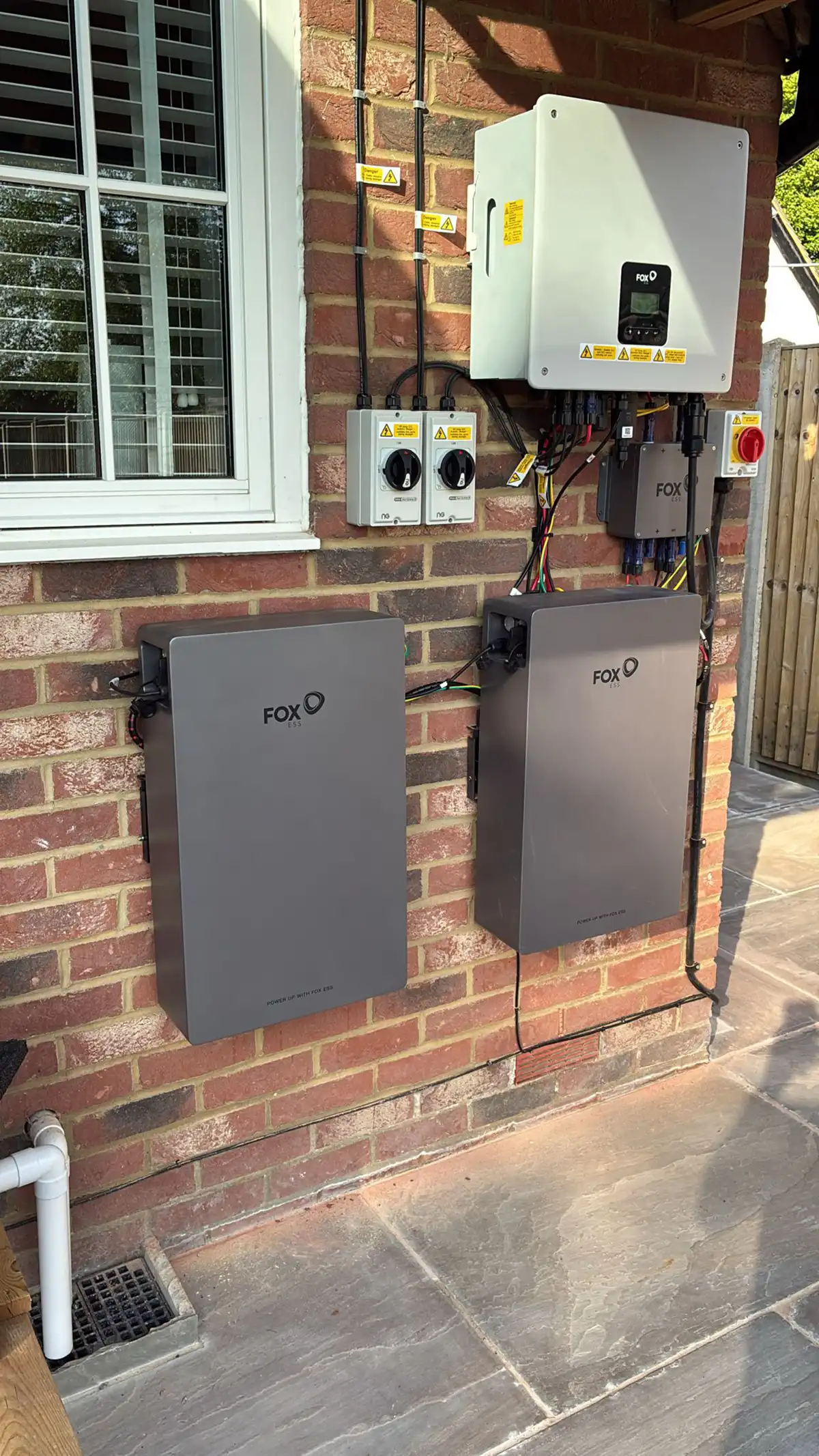 Two FOX-branded battery units and an inverter are mounted on a brick wall beside a window, with various cables and switches connected.