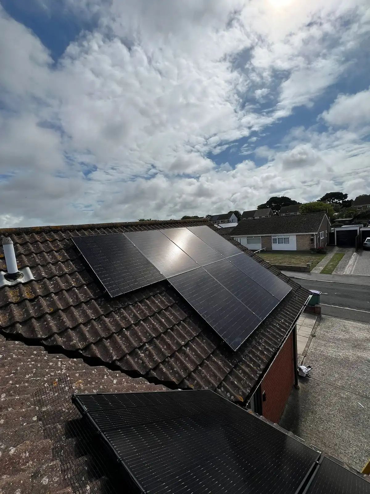 Solar panels installed on the roof of a house under a partly cloudy sky in a residential neighbourhood.