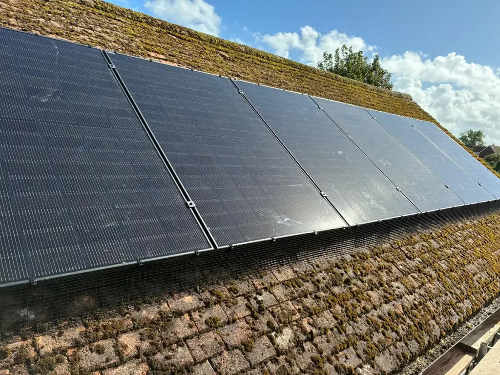 Rows of solar panels installed on a moss-covered, sloping roof under a partly cloudy blue sky.