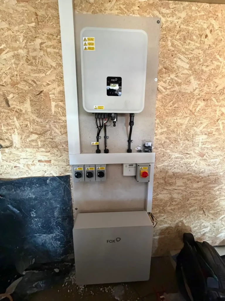 A wall-mounted solar inverter and battery storage unit with cables and switches installed on a wooden panel.