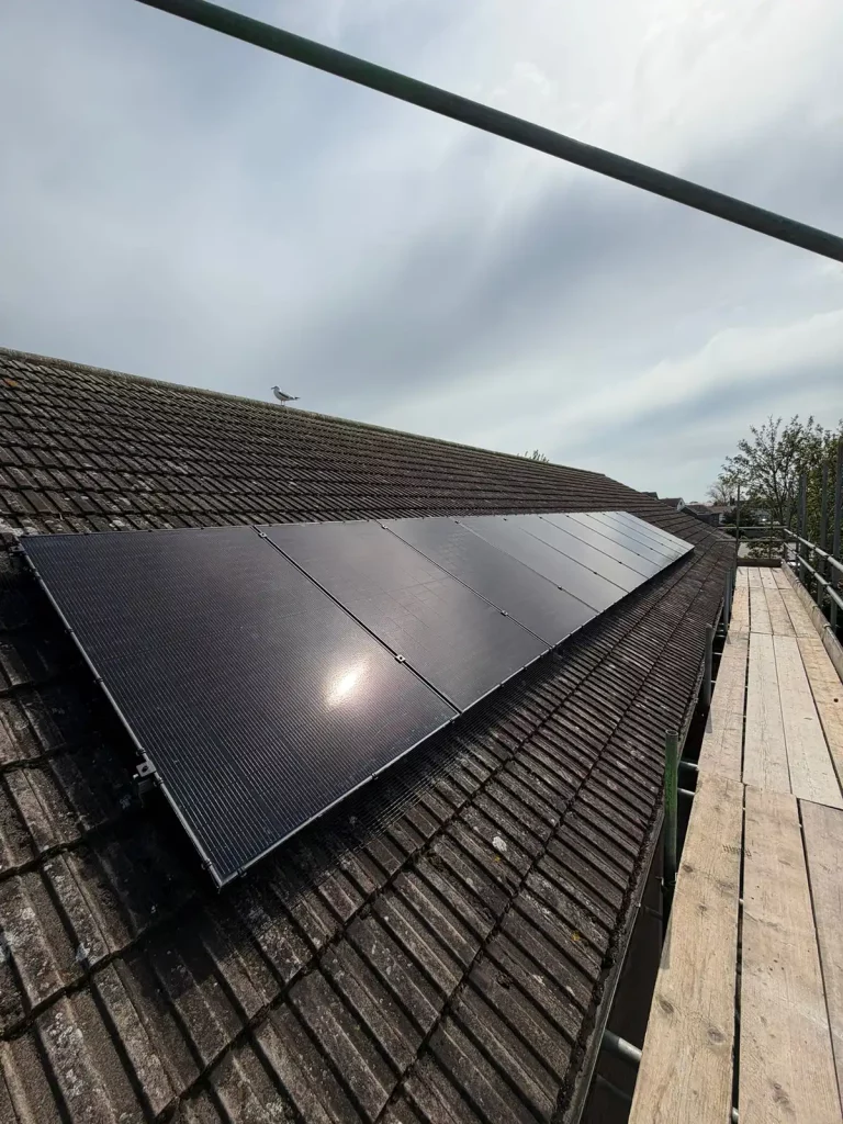 A row of black solar panels installed on a sloping, tiled roof under a partly cloudy sky, with scaffolding at the side.