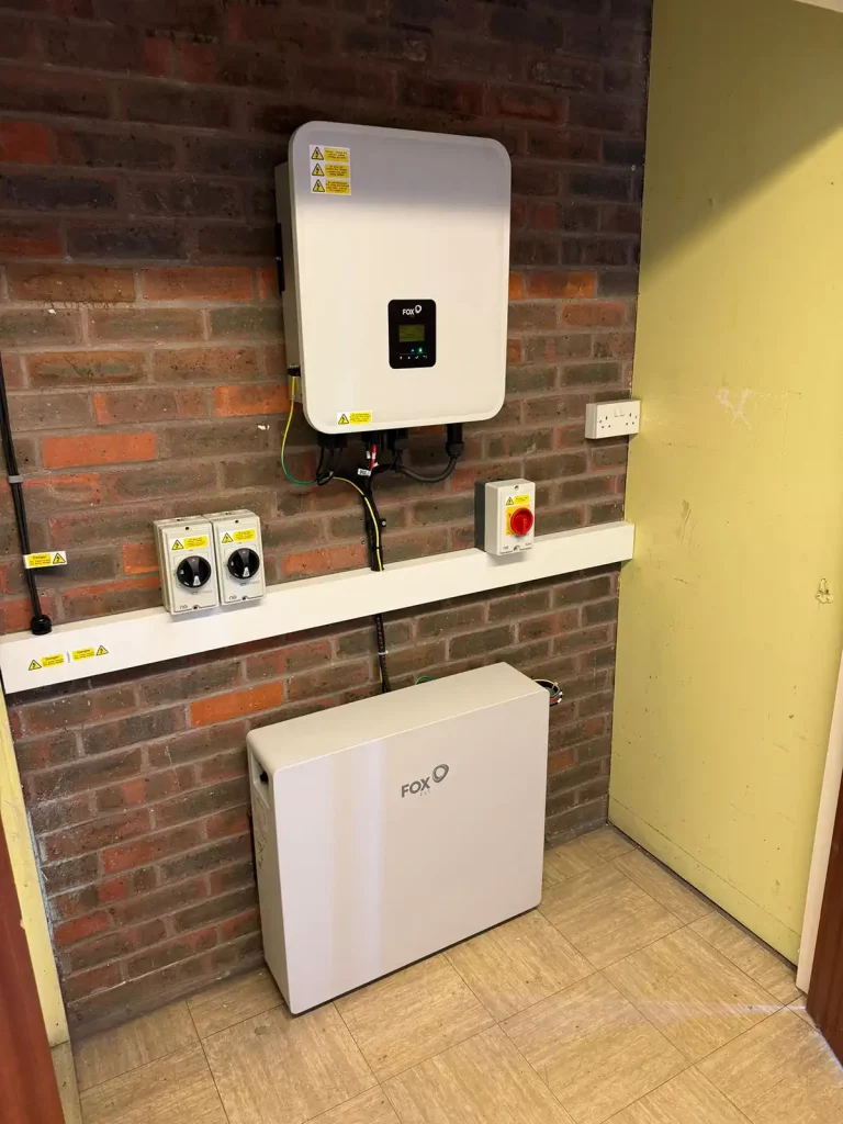 Wall-mounted inverter and control switches above a FOX battery unit in a room with brick walls and tiled flooring.