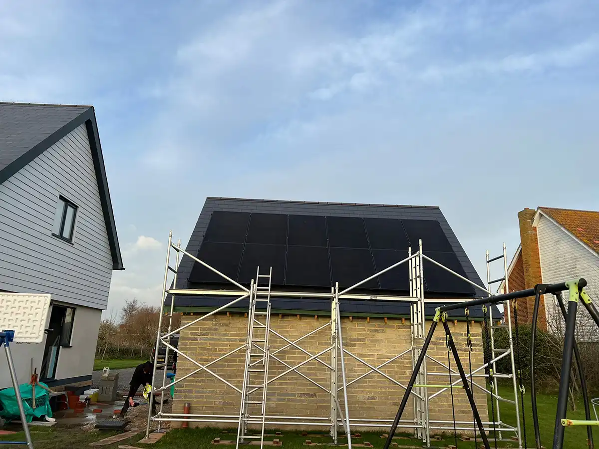 A small brick building with a black solar panel roof, surrounded by scaffolding, stands in a grassy garden under a blue sky.