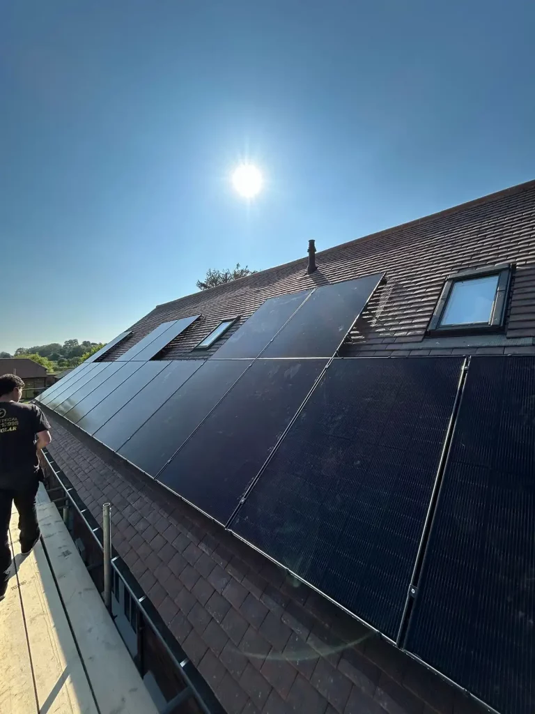Rows of solar panels installed on a sloping roof under a bright sun, with a person standing on scaffolding nearby.
